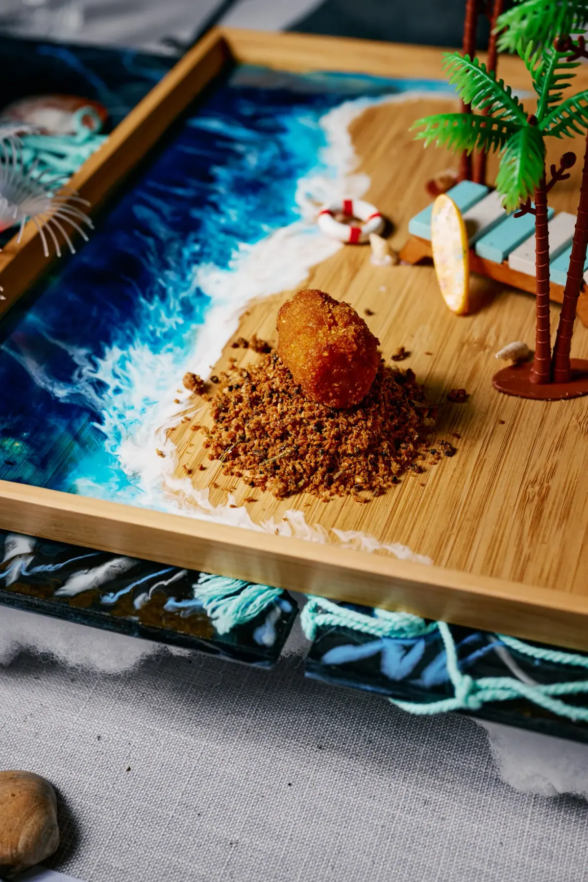 A dessert on a crumble of sand beside a resin wave, served on a wooden board
