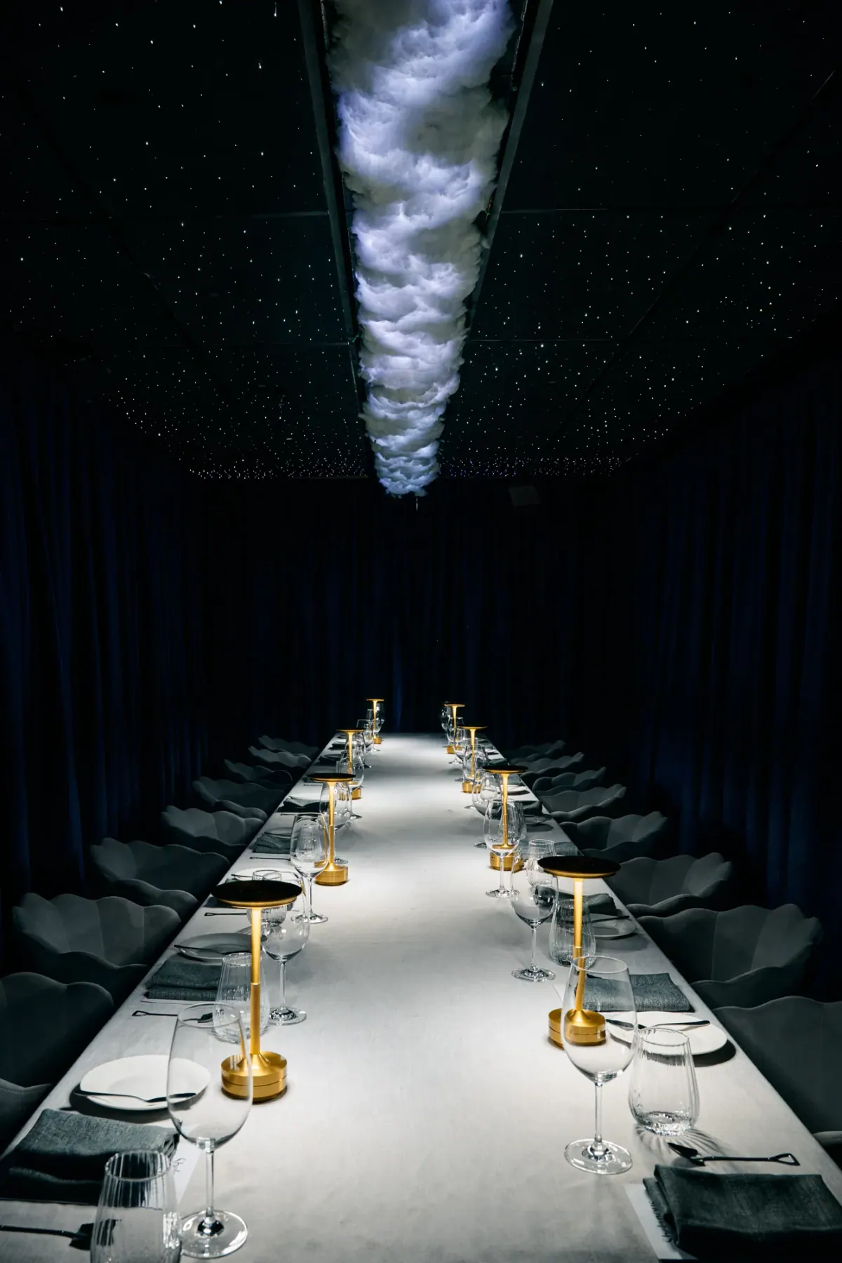 The long table lit along its centre under the cloud, blue curtains all around